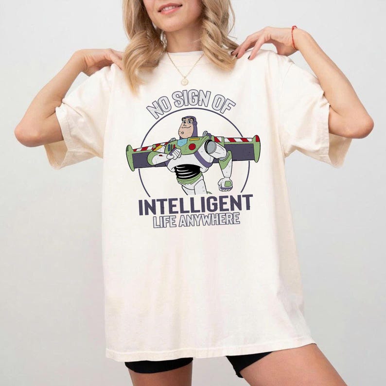 Puede incluir: Camiseta color crema con una imagen de dibujos animados de Buzz Lightyear y el texto "NO SIGN OF INTELLIGENT LIFE ANYWHERE". El dise&ntilde;o incluye las alas y el traje espacial de Buzz Lightyear en un c&iacute;rculo.
