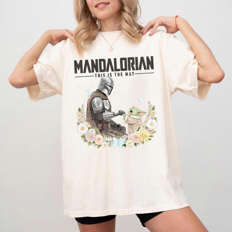 Pode incluir: Camiseta creme com o texto preto "MANDALORIAN - THIS IS THE WAY". Abaixo, um gr&aacute;fico do Mandaloriano e Grogu rodeados por flores e borboletas. A camiseta &eacute; um estilo casual unissex.