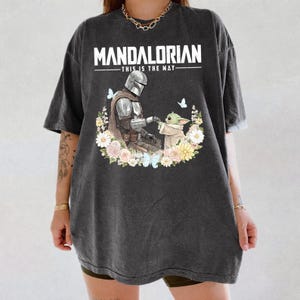 Pode incluir: T-shirt cinzenta escura com um desenho de The Mandalorian e Grogu, rodeados por flores e borboletas. O texto "MANDALORIAN - THIS IS THE WAY" est&aacute; impresso em branco. A t-shirt tem mangas curtas e um corte folgado.