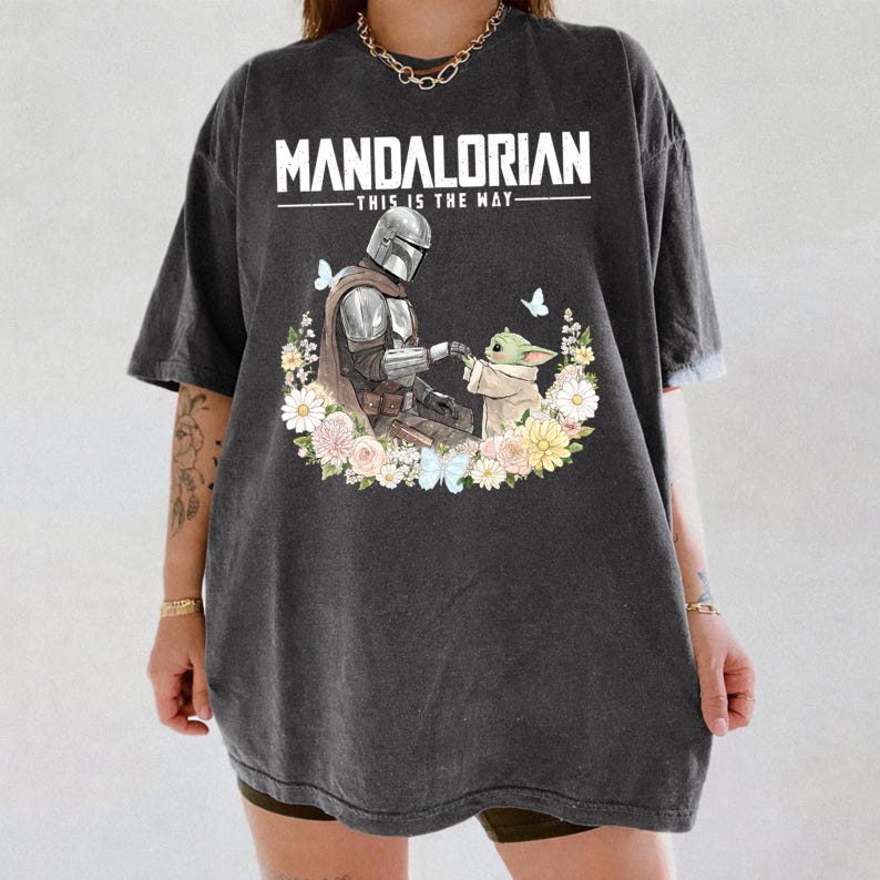 Pode incluir: T-shirt cinzenta escura com um desenho de The Mandalorian e Grogu, rodeados por flores e borboletas. O texto "MANDALORIAN - THIS IS THE WAY" est&aacute; impresso em branco. A t-shirt tem mangas curtas e um corte folgado.