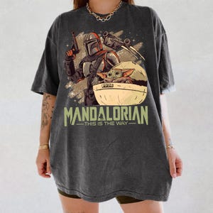 May include: A charcoal gray oversized t-shirt featuring a graphic print of a Mandalorian character holding a blaster, a small green creature in a floating pod, and a spaceship. The text reads MANDALORIAN THIS IS THE WAY in a light green font.