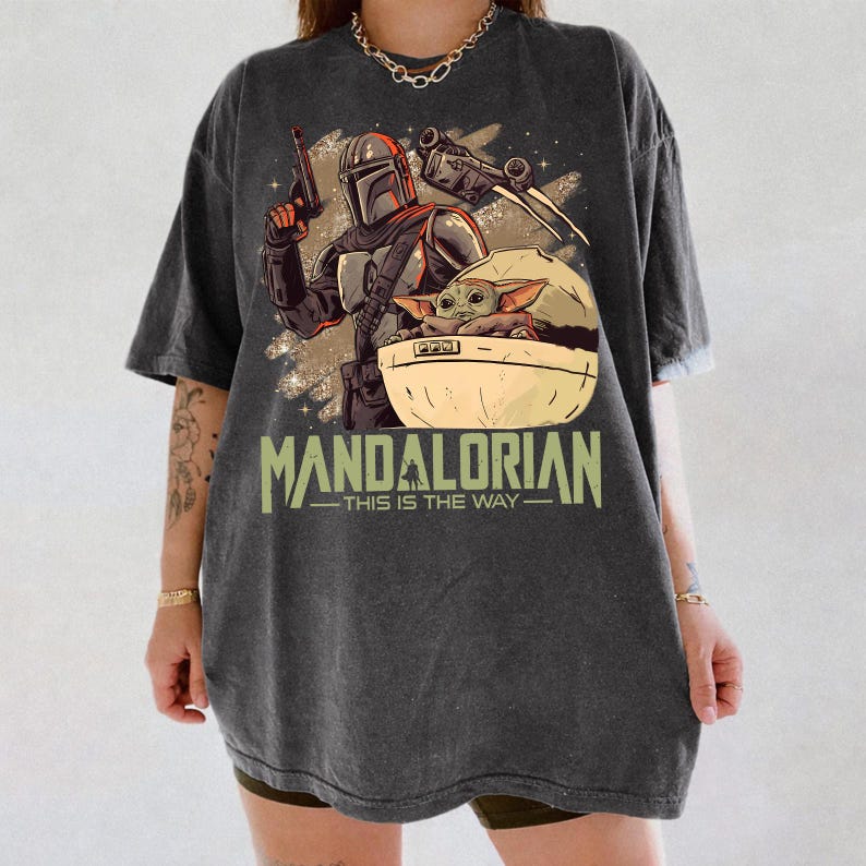 May include: A charcoal gray oversized t-shirt featuring a graphic print of a Mandalorian character holding a blaster, a small green creature in a floating pod, and a spaceship. The text reads MANDALORIAN THIS IS THE WAY in a light green font.