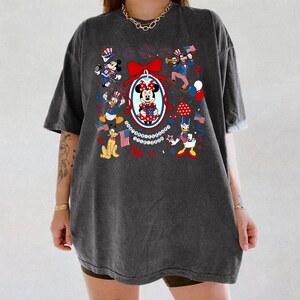 May include: Dark gray t-shirt featuring a patriotic design with Mickey and Minnie Mouse, Donald Duck, Goofy, and other Disney characters. The design includes red, white, and blue elements, flags, and fireworks, celebrating Independence Day.