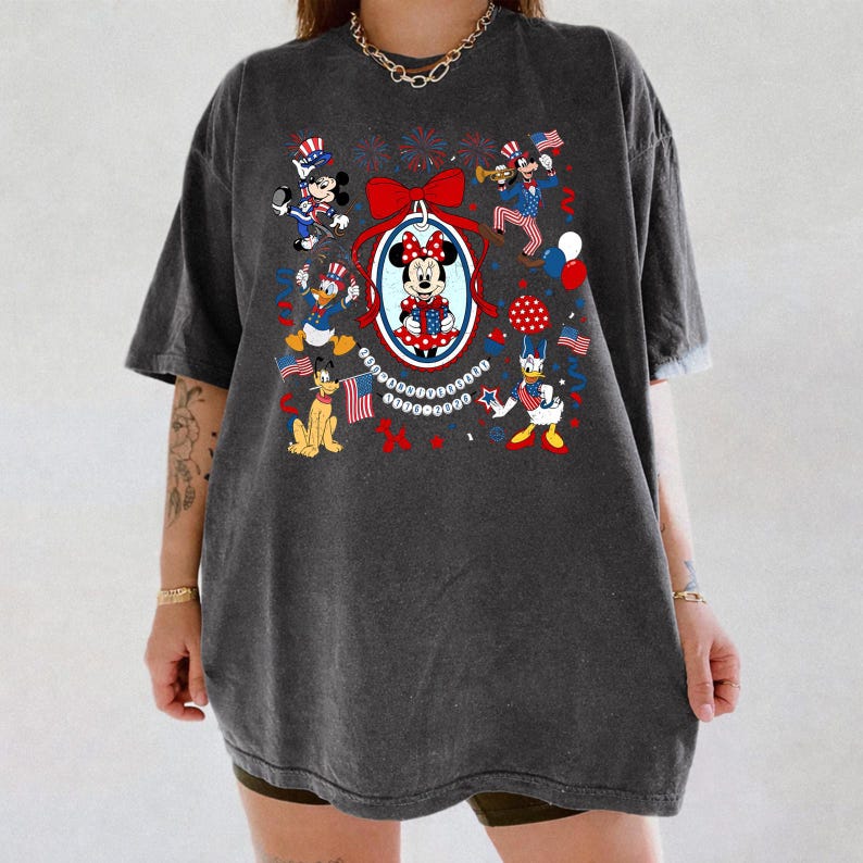 May include: Dark gray t-shirt featuring a patriotic design with Mickey and Minnie Mouse, Donald Duck, Goofy, and other Disney characters. The design includes red, white, and blue elements, flags, and fireworks, celebrating Independence Day.