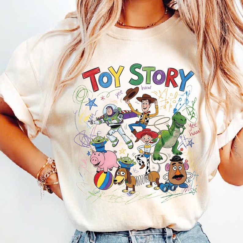 May include: Cream-colored t-shirt featuring the words "TOY STORY" in colorful letters. The shirt showcases a playful design with cartoon characters like Buzz Lightyear, Woody, and Rex, with colorful doodles.