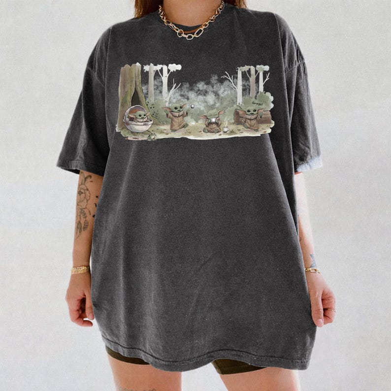 Puede incluir: Camiseta gris oscuro de gran tama&ntilde;o con un gr&aacute;fico de un personaje de una popular franquicia de ciencia ficci&oacute;n. El gr&aacute;fico muestra al personaje en varias poses con un fondo de bosque. El texto "Grogu" tambi&eacute;n es visible.