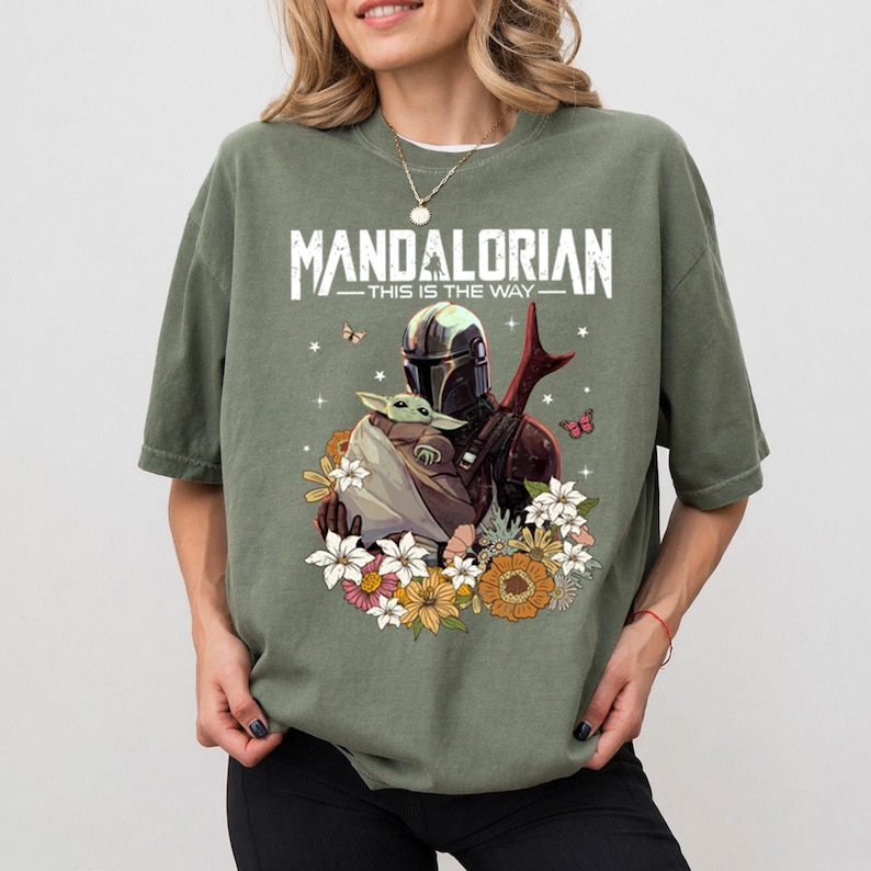 May include: Olive green t-shirt featuring a graphic of The Mandalorian holding Grogu, surrounded by flowers and butterflies. The text "MANDALORIAN - THIS IS THE WAY" is printed across the top in white.