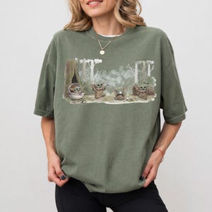 Puede incluir: Camiseta verde oliva con un dise&ntilde;o de Grogu de The Mandalorian. El dise&ntilde;o muestra a Grogu en varias poses en un entorno forestal. La palabra "Grogu" est&aacute; incluida en el dise&ntilde;o.