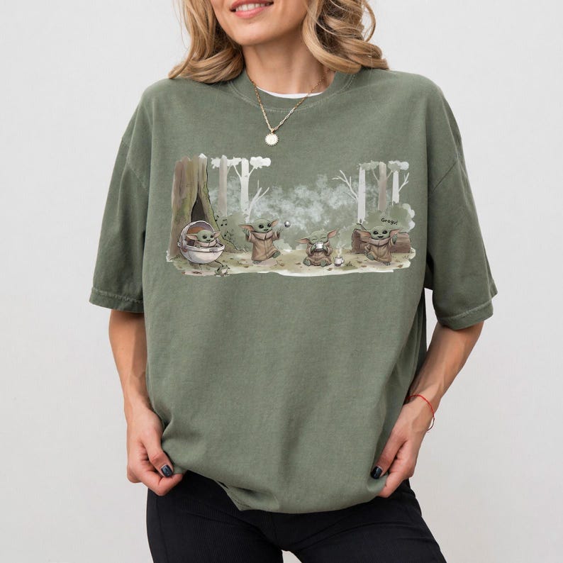 Puede incluir: Camiseta verde oliva con un dise&ntilde;o de Grogu de The Mandalorian. El dise&ntilde;o muestra a Grogu en varias poses en un entorno forestal. La palabra "Grogu" est&aacute; incluida en el dise&ntilde;o.
