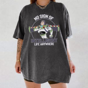 Puede incluir: Camiseta gris oscuro con un dise&ntilde;o de dibujos animados de Buzz Lightyear y el texto "NO SIGN OF INTELLIGENT LIFE ANYWHERE". El dise&ntilde;o est&aacute; en un c&iacute;rculo. La camiseta es de corte holgado.
