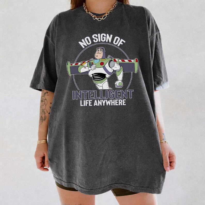 Puede incluir: Camiseta gris oscuro con un dise&ntilde;o de dibujos animados de Buzz Lightyear y el texto "NO SIGN OF INTELLIGENT LIFE ANYWHERE". El dise&ntilde;o est&aacute; en un c&iacute;rculo. La camiseta es de corte holgado.