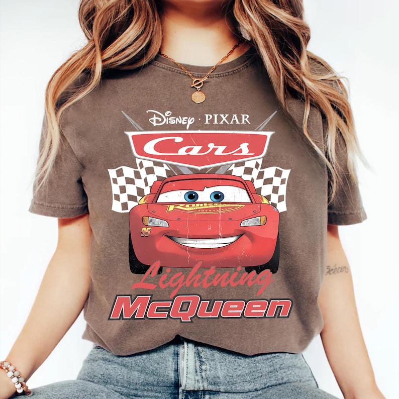 May include: A brown t-shirt featuring a graphic of Lightning McQueen from Disney Pixar's Cars. The design includes the Cars logo, checkered flags, and the text "Lightning McQueen". The shirt has a crew neck and short sleeves.