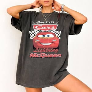 May include: Dark gray t-shirt featuring the Disney Pixar Cars logo and Lightning McQueen. The graphic includes a red race car with the number 95, a checkered flag, and the text "Lightning McQueen" in red.