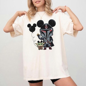 May include: Cream-coloured t-shirt with a cartoon illustration of a Mandalorian character wearing Mickey Mouse ears, holding a balloon. The character is with a Grogu figure. The design is in shades of grey, black, red, and green.