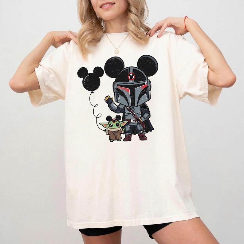 May include: Cream-coloured t-shirt with a cartoon illustration of a Mandalorian character wearing Mickey Mouse ears, holding a balloon. The character is with a Grogu figure. The design is in shades of grey, black, red, and green.