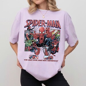 May include: A light purple t-shirt featuring a Spider-Man graphic. The design includes the words "SPIDER-MAN" in red and blue, along with comic book panels and the phrase "WITH GREAT POWER COMES GREAT RESPONSIBILITY."