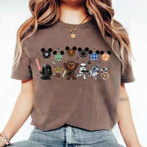 May include: Brown t-shirt featuring a Star Wars-themed design. The design includes cartoon versions of Darth Vader, Yoda, Chewbacca, a Stormtrooper, R2-D2, and BB-8, along with Mickey Mouse ears. The shirt has a crew neck and short sleeves.
