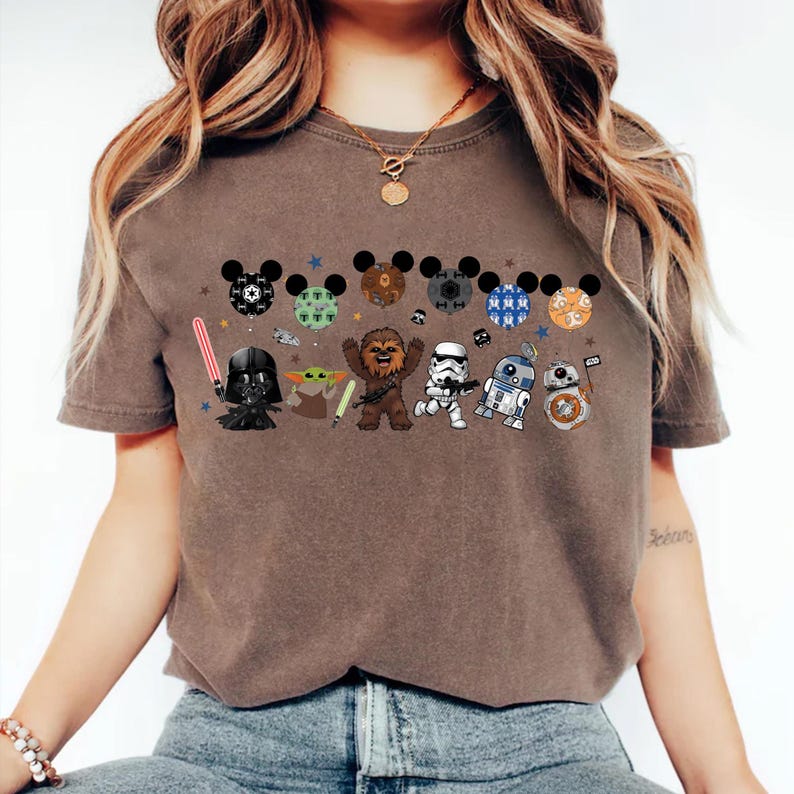 May include: Brown t-shirt featuring a Star Wars-themed design. The design includes cartoon versions of Darth Vader, Yoda, Chewbacca, a Stormtrooper, R2-D2, and BB-8, along with Mickey Mouse ears. The shirt has a crew neck and short sleeves.