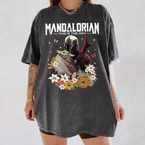 May include: Dark gray t-shirt featuring a graphic of The Mandalorian and Grogu surrounded by flowers and butterflies. The text "MANDALORIAN - THIS IS THE WAY" is printed across the top in white.