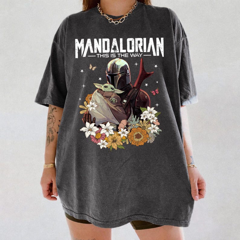 May include: Dark gray t-shirt featuring a graphic of The Mandalorian and Grogu surrounded by flowers and butterflies. The text "MANDALORIAN - THIS IS THE WAY" is printed across the top in white.