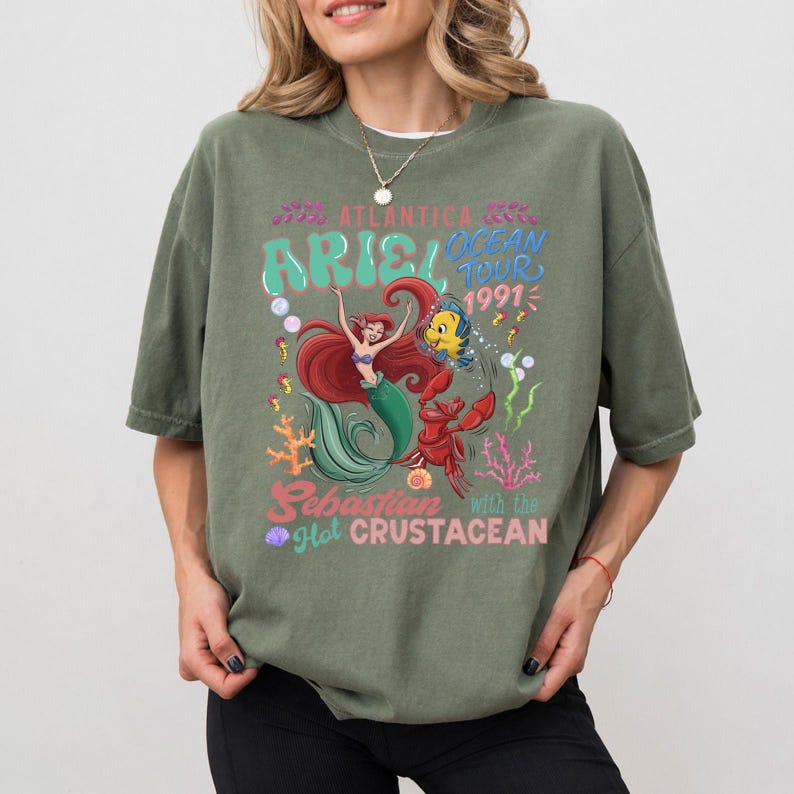 Puede incluir: Camiseta verde oliva con un gr&aacute;fico colorido de Ariel, Sebasti&aacute;n y Flounder de La Sirenita. El dise&ntilde;o incluye el texto "Ariel Ocean Tour 1991" y "Sebastian with the Hot Crustacean."