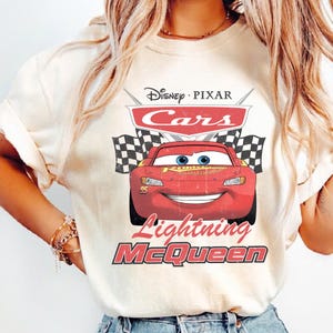 May include: Cream-colored t-shirt featuring the Disney Pixar Cars logo with Lightning McQueen. The design includes a red race car, checkered flags, and the text "Lightning McQueen". The shirt has a classic crew neck.