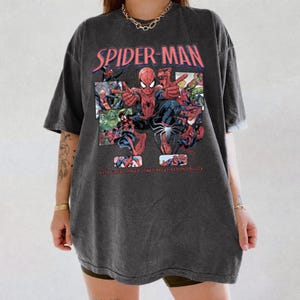 May include: Dark gray t-shirt featuring a Spider-Man graphic. The design includes the words "SPIDER-MAN" in red and a comic-style illustration of Spider-Man and other characters. The text "WITH GREAT POWER COMES GREAT RESPONSIBILITY" is also visible.