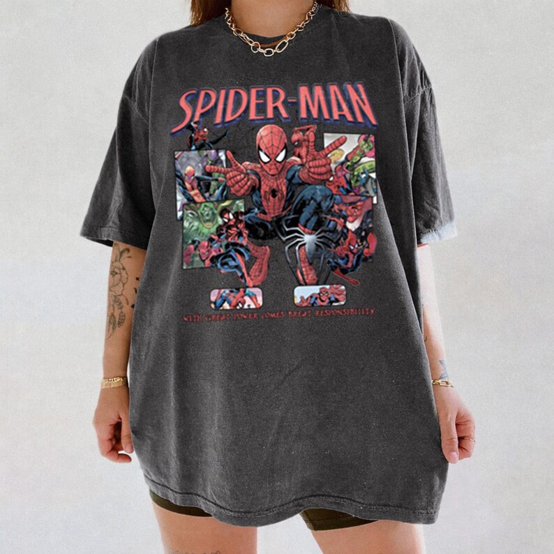 May include: Dark gray t-shirt featuring a Spider-Man graphic. The design includes the words "SPIDER-MAN" in red and a comic-style illustration of Spider-Man and other characters. The text "WITH GREAT POWER COMES GREAT RESPONSIBILITY" is also visible.