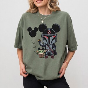 May include: An olive green t-shirt with a cartoon illustration of a Mandalorian character with Mickey Mouse ears, holding a black balloon. A Grogu character is next to the Mandalorian. The t-shirt has a loose fit.