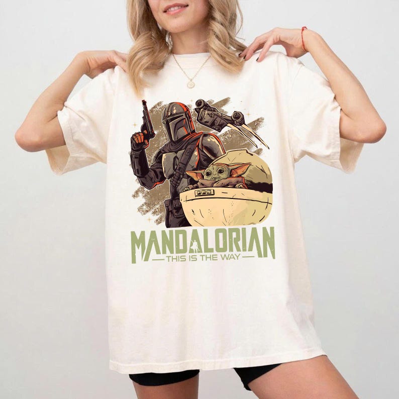 May include: An off-white t-shirt featuring a graphic print of a Mandalorian character holding a blaster, a small green creature in a floating pod, and the text "MANDALORIAN" above the phrase "THIS IS THE WAY" in a muted green font.