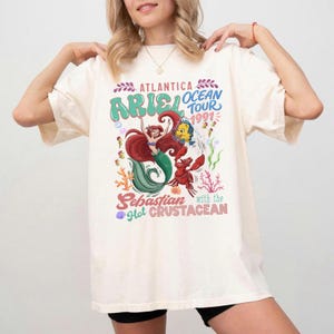 Puede incluir: Camiseta color crema con un gr&aacute;fico colorido de Ariel, Flounder y Sebasti&aacute;n de La Sirenita. El dise&ntilde;o incluye el texto "Atlantica Ocean Tour 1991" y "Sebastian with the Hot Crustacean."