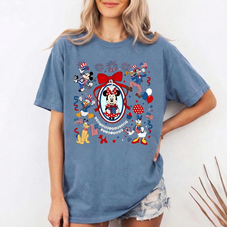 May include: A blue t-shirt featuring a patriotic design with cartoon characters. The graphic includes Mickey and Minnie Mouse, Donald Duck, Pluto, and Goofy, all in red, white, and blue attire, celebrating with flags and fireworks.