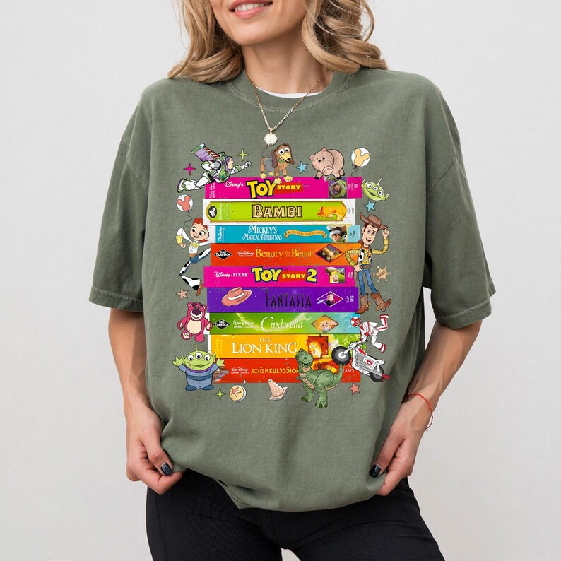 May include: An olive green t-shirt featuring a stack of colorful Disney movie titles and characters. The titles include "Toy Story," "Bambi," "Beauty and the Beast," and "The Lion King." Various cartoon characters are also displayed.