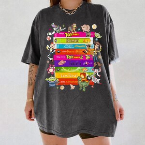 May include: Dark gray t-shirt featuring a stack of Disney movie titles including "Toy Story," "Bambi," and "The Lion King." The design includes colorful book spines and character illustrations from the movies.