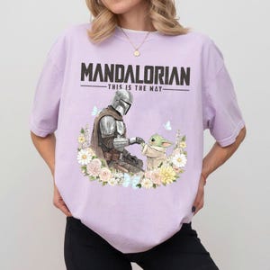 Pode incluir: Camiseta lil&aacute;s clara com um desenho do Mandaloriano e Grogu rodeados por flores e borboletas. O texto "MANDALORIAN - THIS IS THE WAY" est&aacute; impresso em letras pretas na parte superior.