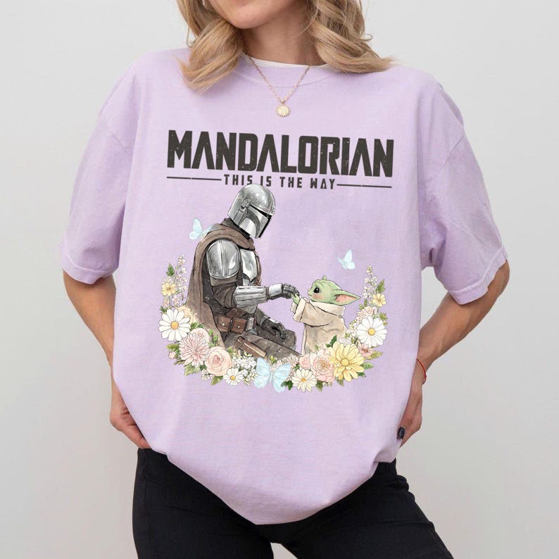 Pode incluir: Camiseta lil&aacute;s clara com um desenho do Mandaloriano e Grogu rodeados por flores e borboletas. O texto "MANDALORIAN - THIS IS THE WAY" est&aacute; impresso em letras pretas na parte superior.