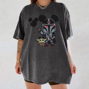 May include: Dark grey t-shirt with a cartoon Mandalorian character wearing Mickey Mouse ears, holding a black balloon. A Grogu figure stands beside the character. The design is printed on a casual, oversized tee.