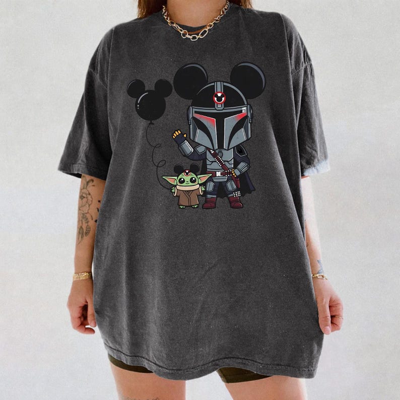 May include: Dark grey t-shirt with a cartoon Mandalorian character wearing Mickey Mouse ears, holding a black balloon. A Grogu figure stands beside the character. The design is printed on a casual, oversized tee.