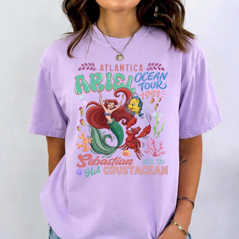 Puede incluir: Una camiseta de color lila claro con un gr&aacute;fico colorido de Ariel, Sebasti&aacute;n y Flounder de La Sirenita. El dise&ntilde;o incluye el texto "Atlantica Ocean Tour 1991" y "Sebastian with the Hot Crustacean."