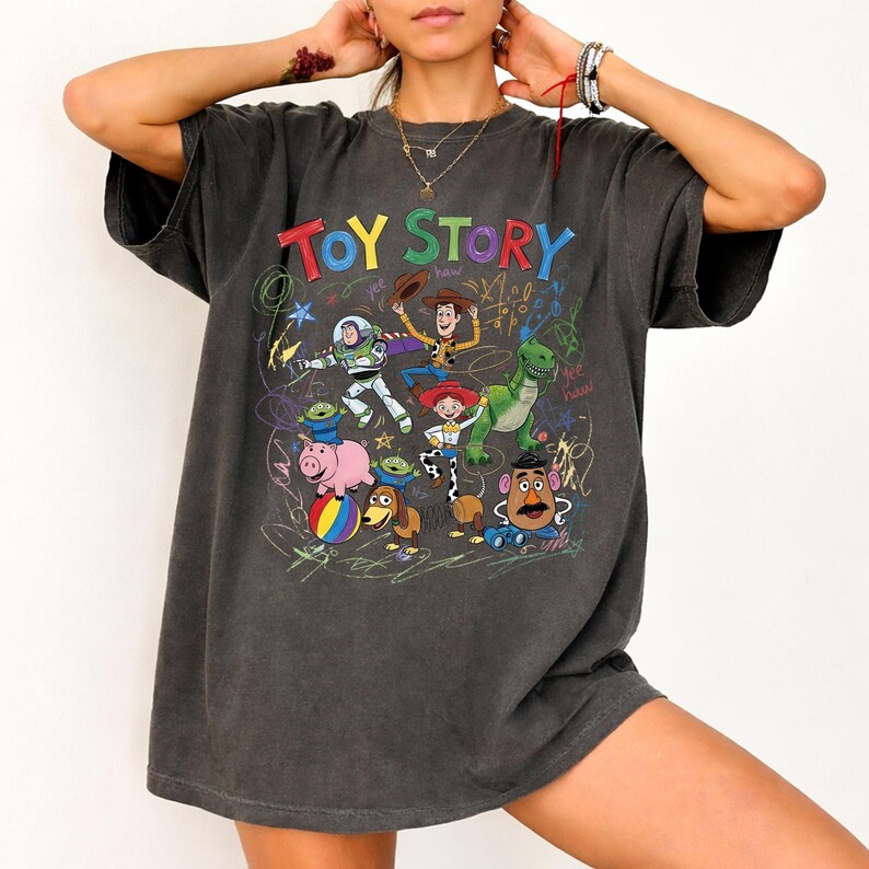 May include: Dark gray t-shirt featuring the words "TOY STORY" in colorful letters above a graphic of characters from the animated film. The design includes Woody, Buzz Lightyear, Rex, and other characters. The shirt has a vintage, worn look.