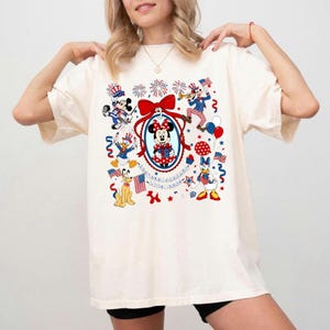 May include: Cream-colored t-shirt featuring a patriotic Minnie Mouse design. The design includes Minnie Mouse in a red, white, and blue dress, surrounded by other Disney characters, fireworks, and American flags. The shirt has a relaxed fit.