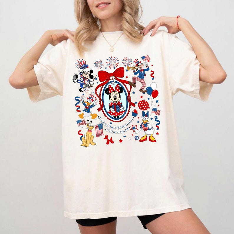 May include: Cream-colored t-shirt featuring a patriotic Minnie Mouse design. The design includes Minnie Mouse in a red, white, and blue dress, surrounded by other Disney characters, fireworks, and American flags. The shirt has a relaxed fit.