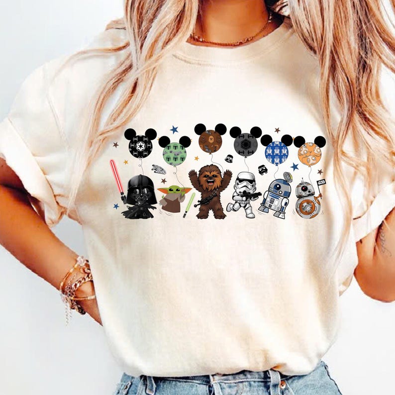 May include: Cream-colored t-shirt featuring a line of Star Wars characters holding Mickey Mouse-shaped balloons. Darth Vader, Yoda, Chewbacca, a Stormtrooper, R2-D2, and BB-8 are depicted. The balloons have various patterns and colors.