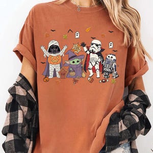 Op de afbeelding: Een roestkleurig t-shirt met een grafische print van vier personages in Halloween-kostuums. De personages zijn een mummie, een heks, een vampier en een robot, omringd door kleine pompoenen, spookjes, vleermuizen en herfstbladeren.