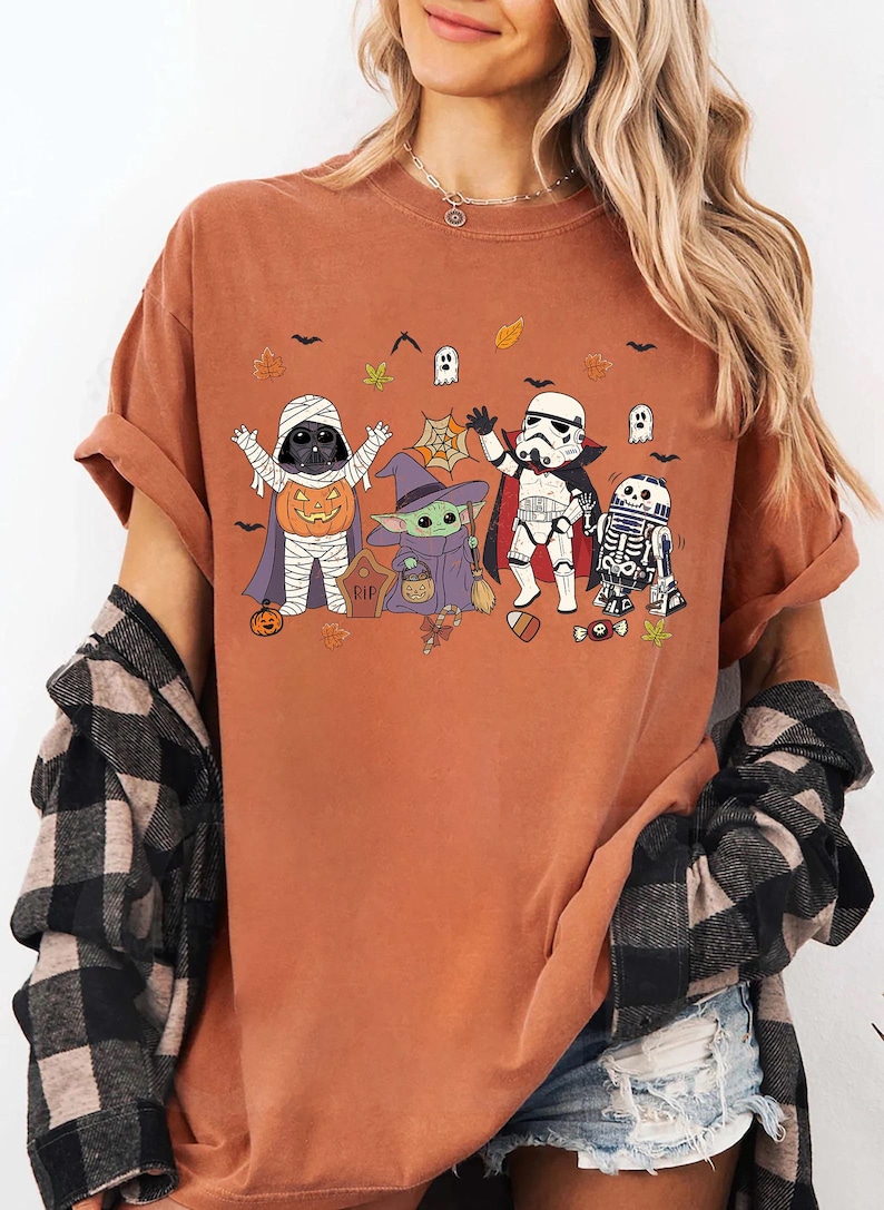 Op de afbeelding: Een roestkleurig t-shirt met een grafische print van vier personages in Halloween-kostuums. De personages zijn een mummie, een heks, een vampier en een robot, omringd door kleine pompoenen, spookjes, vleermuizen en herfstbladeren.