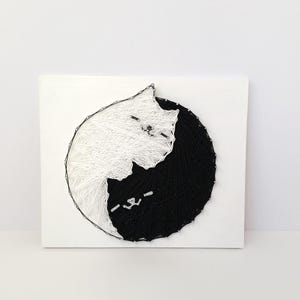 May include: A handmade string art piece featuring a black and white cat design. The cat is depicted in a yin-yang style, with one half in white and the other in black. The artwork is mounted on a white square wooden board.