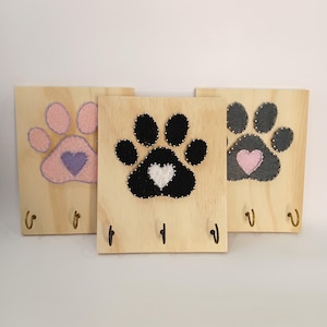 Puede incluir: Tres llaveros de madera con diseños de huellas de patas. Cada soporte presenta una huella de pata de diferente color: rosa, negro y gris. Cada huella de pata tiene un corazón en el centro. Los llaveros tienen tres ganchos de metal.