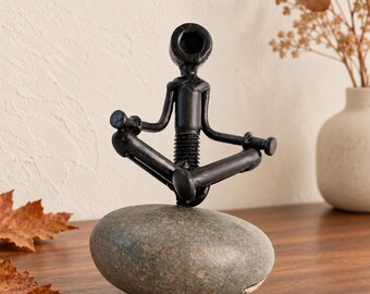May include: A black metal figurine constructed from hardware components like nuts, bolts, and screws, shaped into a person sitting in a cross-legged meditation pose. The figure is securely mounted on top of a smooth, oval-shaped gray river stone.