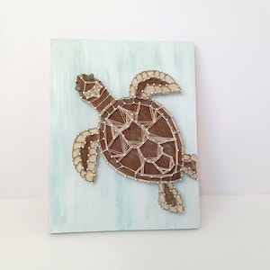 Puede incluir: Una pieza de arte de hilo hecha a mano con un diseño de tortuga marina. La tortuga es marrón con detalles blancos, sobre un fondo de madera pintada de azul claro. Un artículo decorativo único.