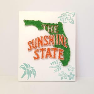 May include: A white canvas with a green string art outline of Florida and the words "THE SUNSHINE STATE" in orange string. Light blue floral designs are in the corners. A unique piece of wall art.